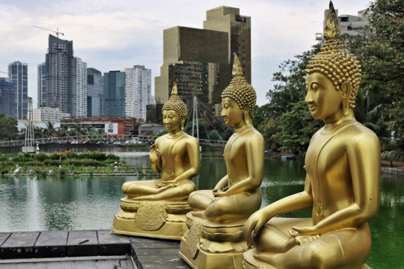 three gold buddha statues in sitting position on a stone wall next to a lake with skyscrapers in the background - taken in Colombo on a 2 weeks Sri Lanka itinerary