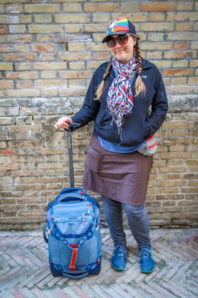 A woman with baseball cap sunglasses and hair in plaits standing next to a small blue cabin bag