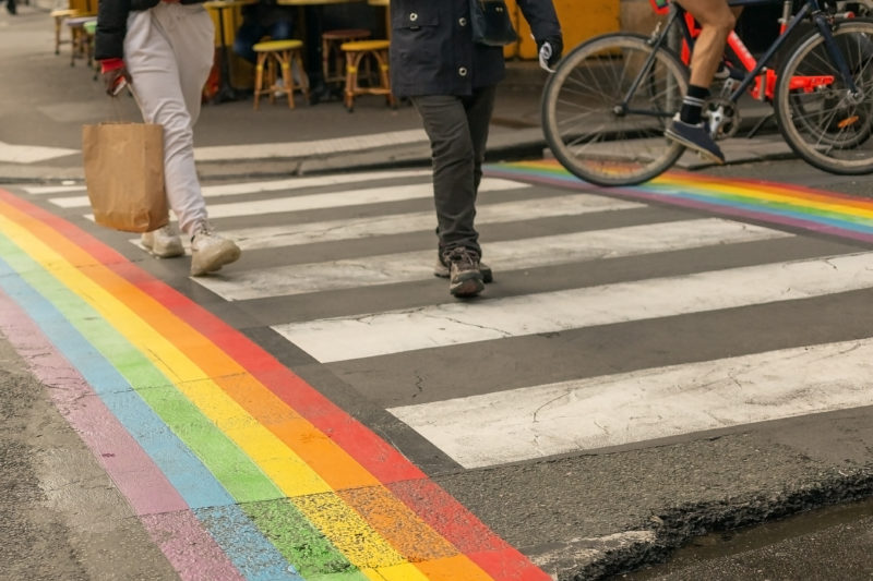 Gay pride flag, on crosswalk with people walking across it in Paris.