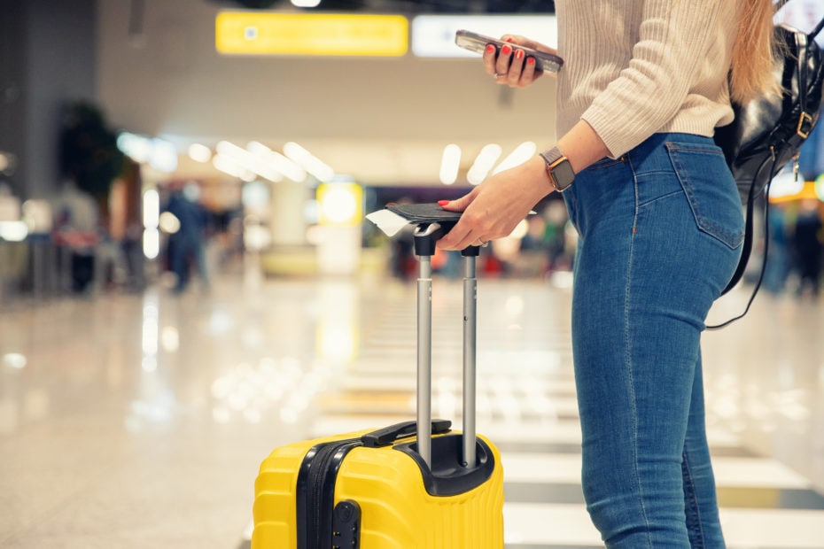 lower half of a woman in jeans holding a yellow suitcase and plane ticket in an airport - best carry on luggage for women