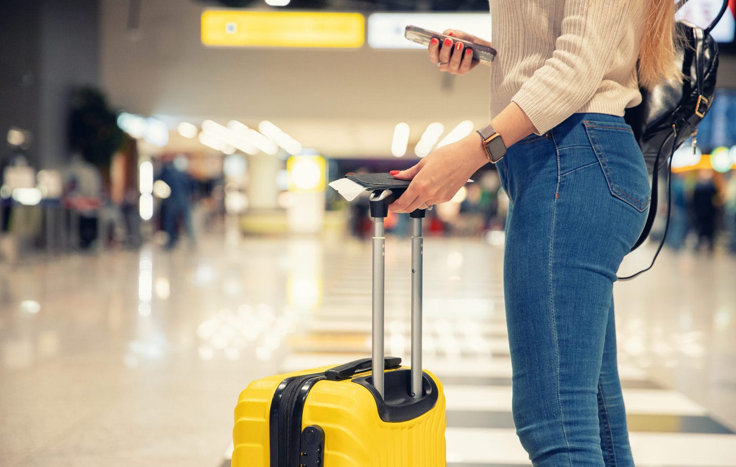 lower half od a woman in jeans holding a yellow suitcase and plane ticket in an airport - best carry on luggage for women