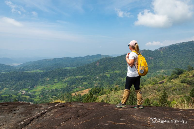 emily wearing a white t shirt, black leggings and brown leech socks with a pink cap and yellow backpack, standing on the edge of a rock cliff edge looking out at a lush green valley below while hiking in the knuckles on a 2 weeks sri lanka itinerary