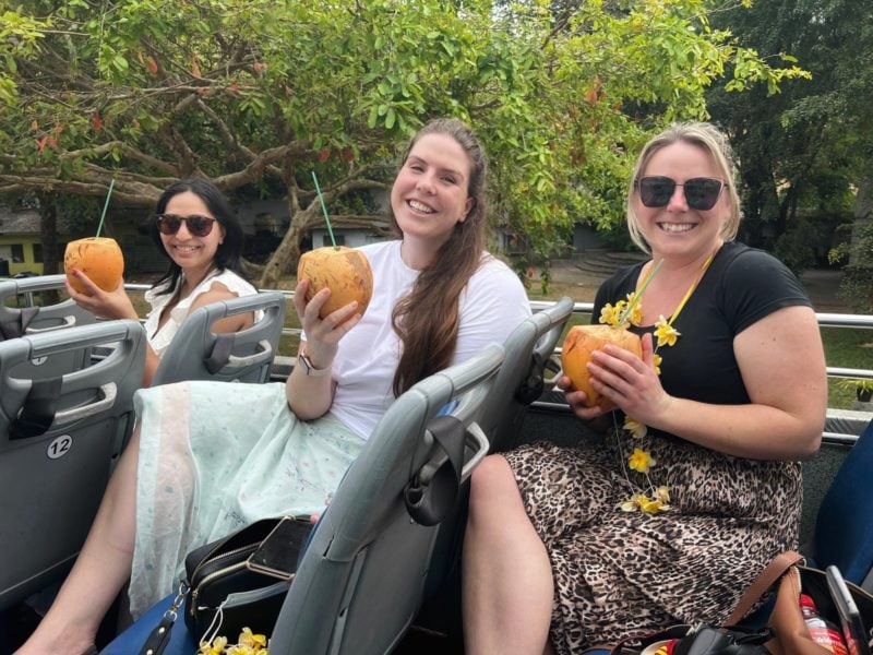 Emily wearing black t-shirt and leopard print skirt holding a coconut, sitting on an open top bus next to Lisa and Roshni who are both wearing white tops and holding coconuts