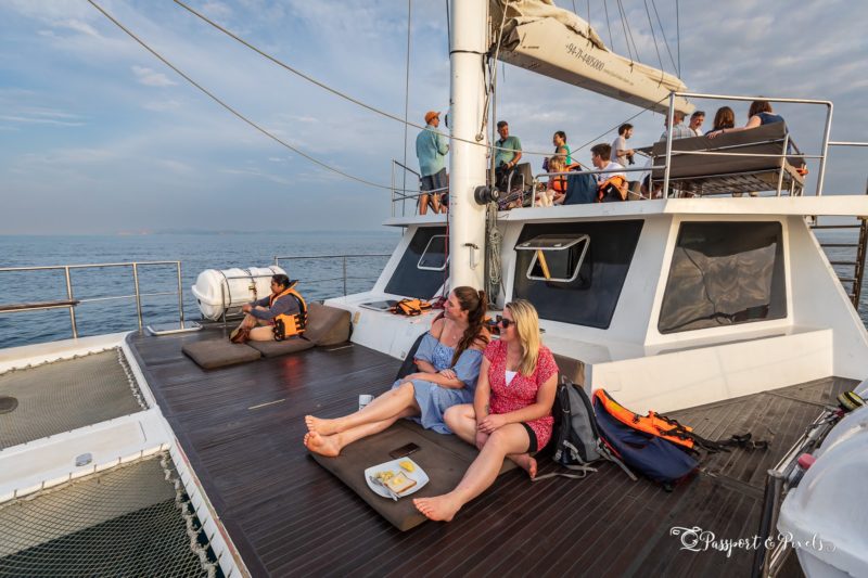 Emily in a red dress sitting on the deck of a yacht next to Lisa in a blue dress, both looking out to sea