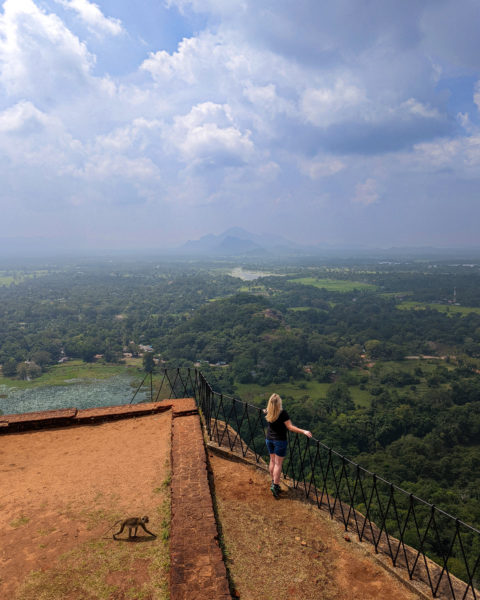 Emily wearing a black top and denim shorts standing on a viewing platform looking out at a view of a green valley far below and mountains in the distance. Taken in Sigiriya on a 2 weeks sri lanka itinerary. 