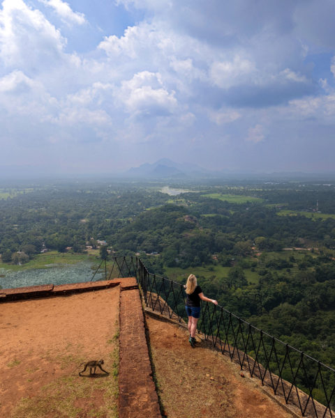 Emily wearing a black top and denim shorts standing on a viewing platform looking out at a view of a green valley far below and mountains in the distance. Taken in Sigiriya on a 2 weeks sri lanka itinerary.
