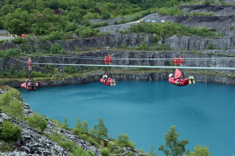 3 people in red jumpsuits and white helmets descended face down from a zip line with red metal hooks fixing them to the wire above a vivid blue lake 
