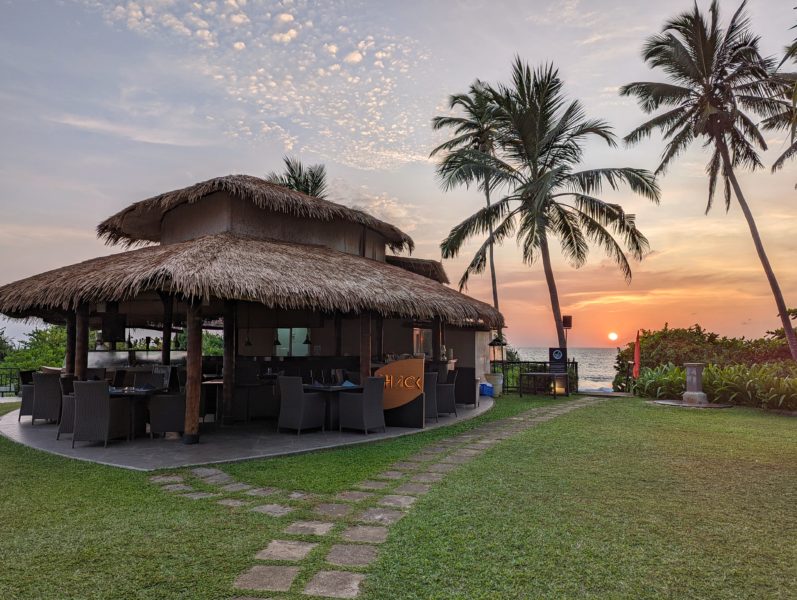 small wooden building with a palm frond roof next to some palm trees with the sun setting over the sea in the background - 2 weeks in sri lanka itinerary