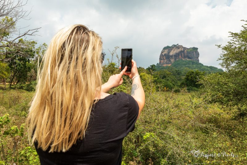 a girl with messy blonde hair taking a photo on her phone of a large rock monolith in the distance at sigiriya in sri lanka