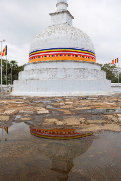 a white stupa with an orange and yellow patterned sash around it reflected in a puddle on a stone floor at Anuradhapura on a 2 weeks sri lanka itinerary