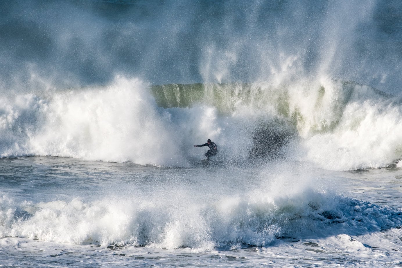a surfer in a black wetsuit in front or a huge crashing wave