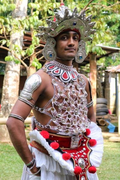 a man in a traditional kandyan dance costume with an elaborate silver headress and a top made of strings of small beads