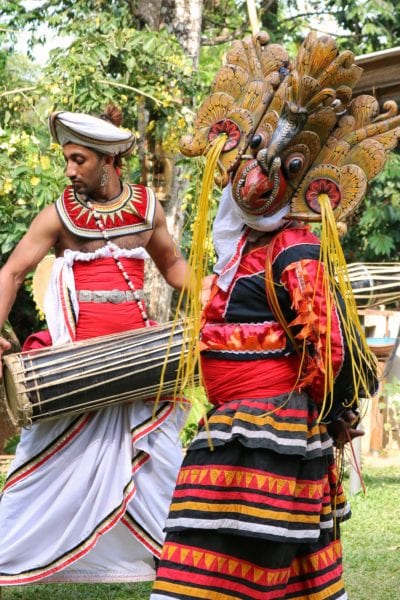 a man in a red costume with  large wooden mask shaped like a bird, dancing in front of a man playing a traditional wooden drum 