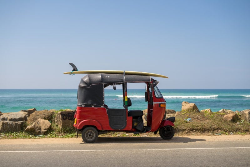 a red tuk tuk parked by the side of a road in front of the ocean in sri lanka with a surfboard attached to its roof. 