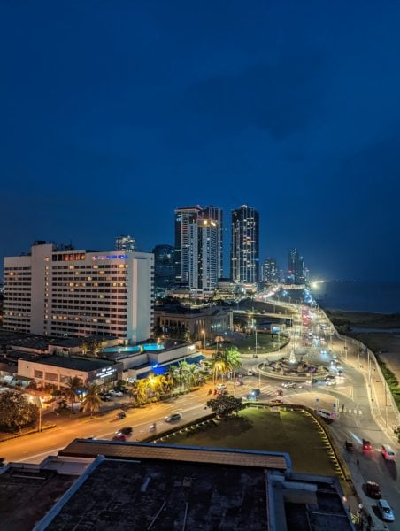 looking down at a busy seafront road in Colombo at night with several skyscrapers lit up in the background