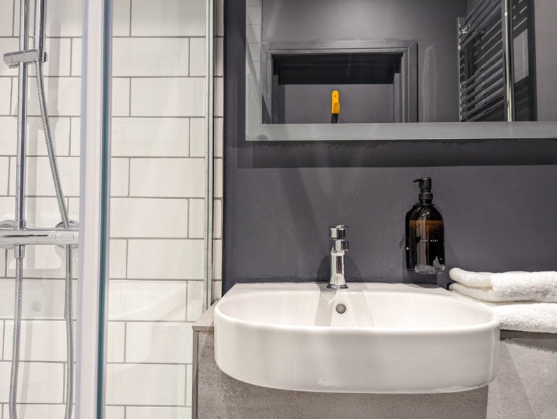 bathroom sink on a grey wall with a mirror above reflecting a grey door and yellow hook