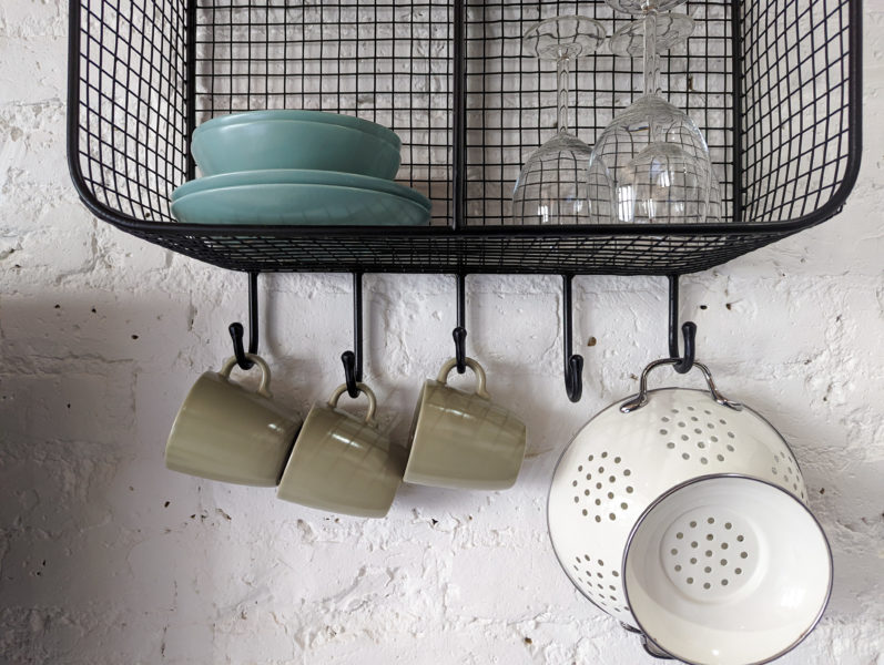 white stone wall with a black wall shelf unit made of wire with some blue bowls and plates on one shelf and wine glasses on another with 3 green cups and a white collander hanging from hooks under the unit