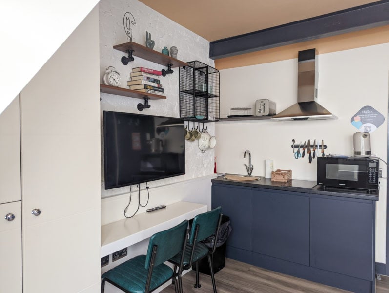 corner of a small studio apartment in south london with a kitchenette with blue-grey cupboards and a wall mountain flat screen tv on a white stone wall