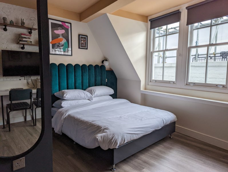bed with green velvet headboard and white duvet in a small serviced apartment in brixton london with wooden floor. there is a mirror in the foreground reflecting shelves and a tv on the opposite wall. 