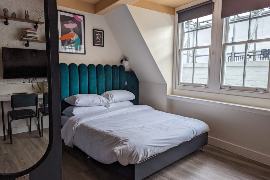 bed with gren velvet headboard and white duvet in a small serviced apartment in brixton london with wooden floor. there is a mirror in the foreground reflecting shelves and a tv on the opposite wall.