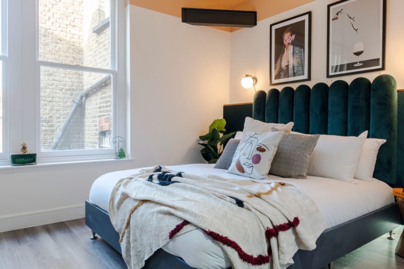 a bed next to a window with white duvet and a colourful throw and a green velvet headboard in a small serviced apartment in brixton london with wooden floor. 