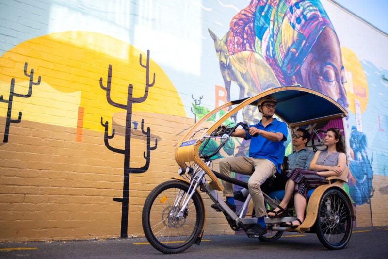 man and woman in a tuk tuk being ridden by an asian man wearing a yellow helmet in front of a large colourful mural on a wall in adelaide
