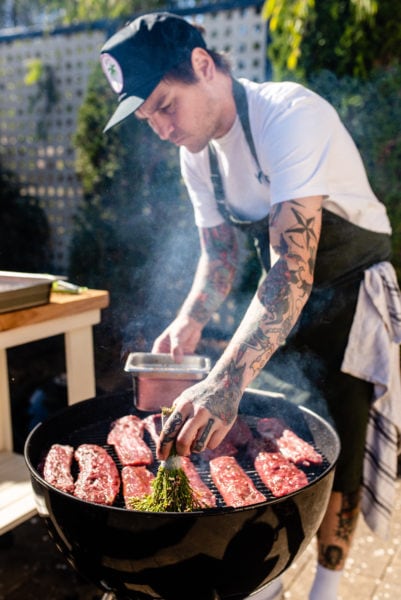man with tattoos and a baseball cap brushing meat grilling on a barbecue