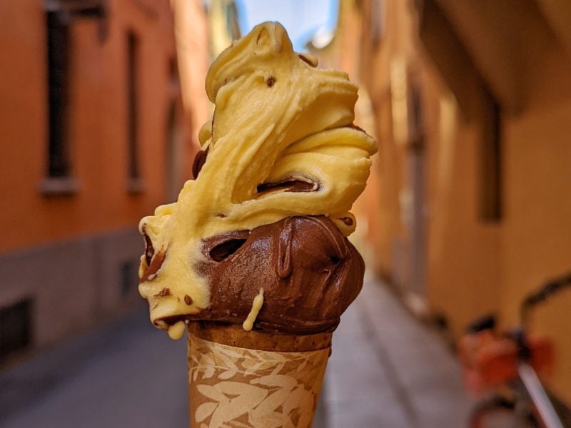 close up of an ice cream cone with a big swirl of yellow custard flavour and brown chocolate flavour gelato in front of a narrow cobbled street in bologna with orange painted walls
