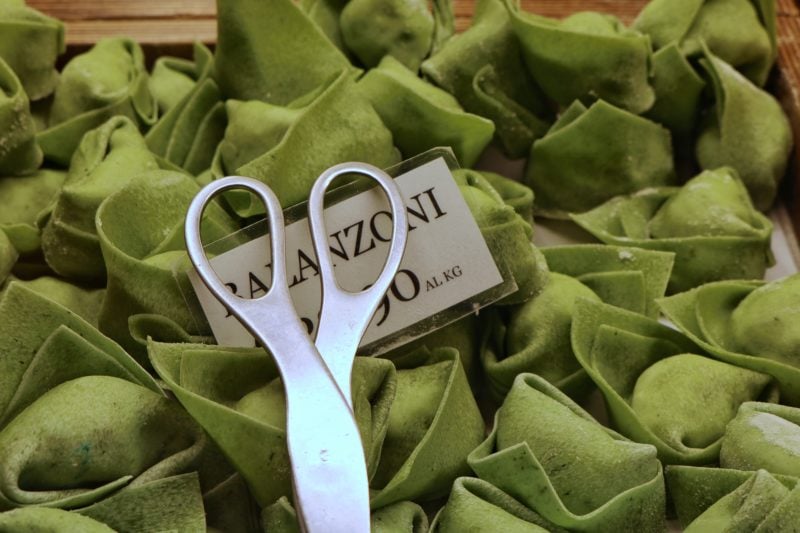 close up of many green coloured stuffed pasta shapes with some metal tongs and a paper sign which says balanzoni with the price obscured underneath. balanzoni pasta are a traditional food from bologna  italy