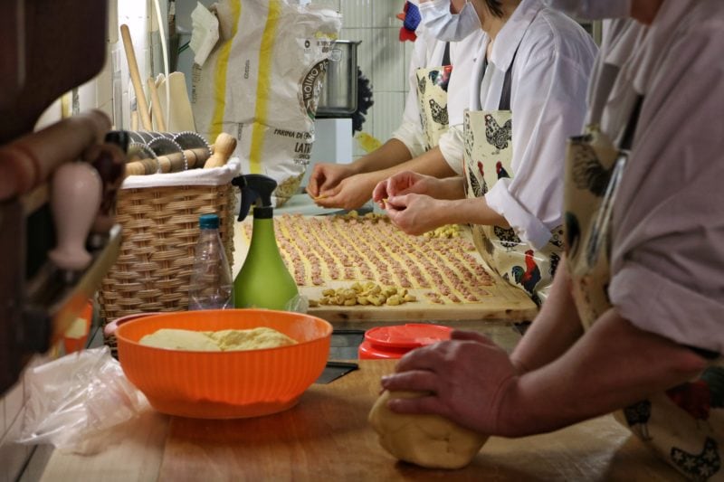 close up of three women's hands making pasta in a shop in bologna italy - the one in the foreground is rolling a large ball of dough while the two behind are filling a large board of pasta with ground meat