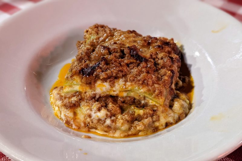 close up of a lasagna on a white plate, with layers of pasta and creamy sauce topped with ground meat in a dark red sauce 