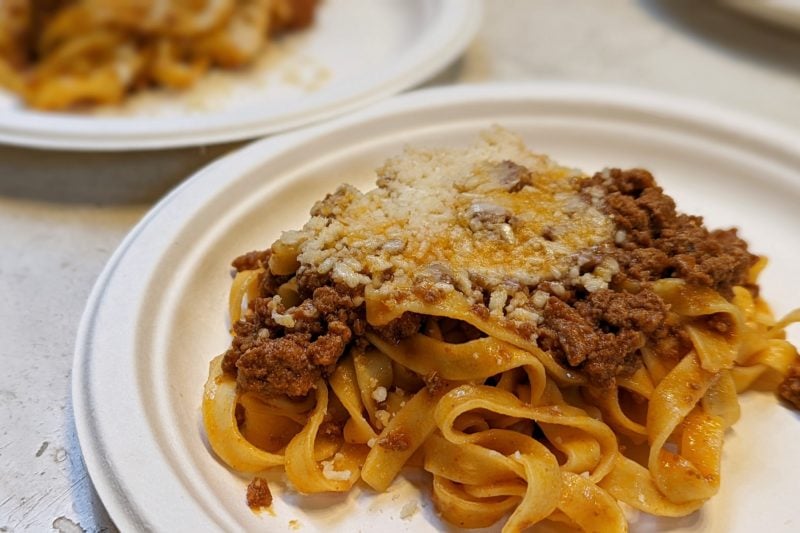 close up of a white paper plate with thick tagliatelle topped with ground meat in a dark red sauce and yellow parmesan shavings served at a food court in bologna italy