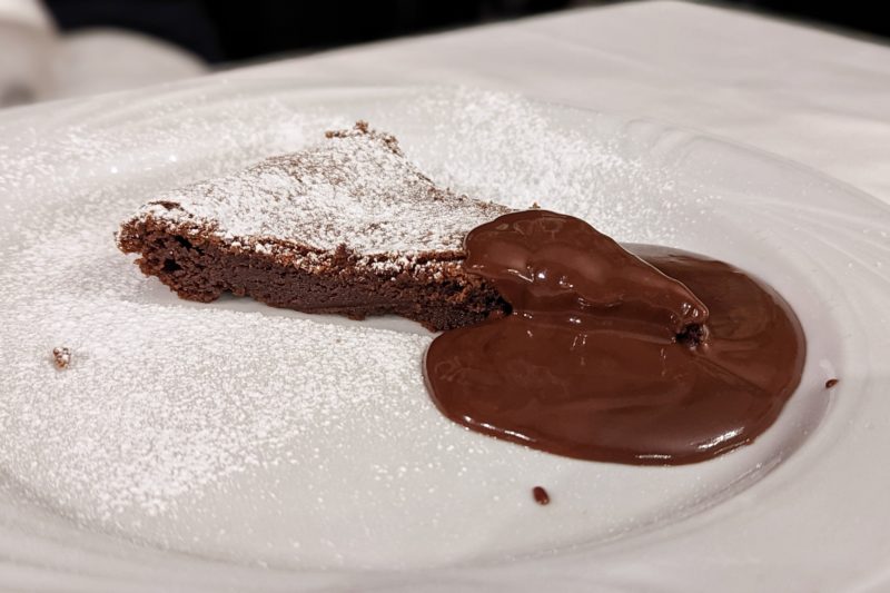 close up of a white plate with a slim triangular slice of chocolate cake next to a large pool of chocolate sauce, all dusted with white powdered sugar - a traditional dessert food in bolgona italy 