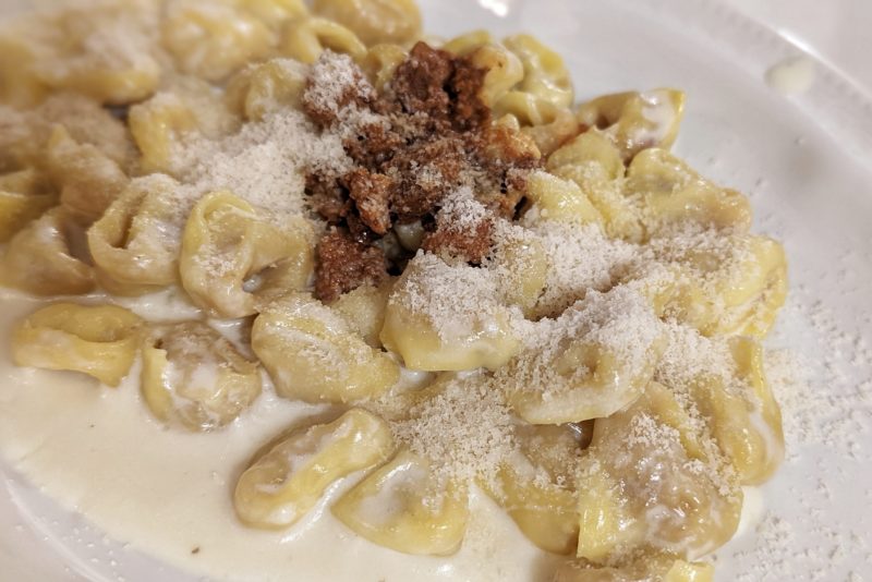 close up of a white plate with small stuffed pasta dumplings in a white sauce topped with a tiny amount of ground meat in a red rag sauce and lots of grated parmesan