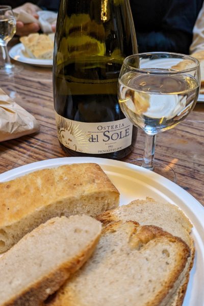 white paper plate with several pieces of crusty bread with a small wine glass behind it filled with an almost clear sparkling wine next to a dark green glass wine bottle with the label reading osteria del sole