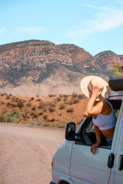 girl leaning out of a van window and holding her floppy hat on a dirt road in front of an orange rocy mountain. working holiday visa in adelaide and south australia. 