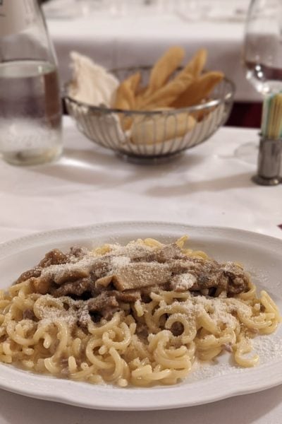 a white plate with small curled pasta shapes in a white sauce topped with pieces of ground sausage meat and sliced mushrooms and parmesan with a white tablecloth in the background and a silver dish containing pieces of bread at a restaurant in bologna italy