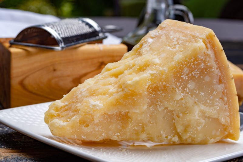 close up of a large triangular chunk of crumbly pale yellow parmesan cheese with a metal grater and wooden chopping board behind it - bologna food guide