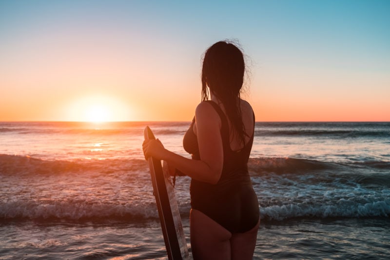 girl with a surf board in front of sunset over the sea