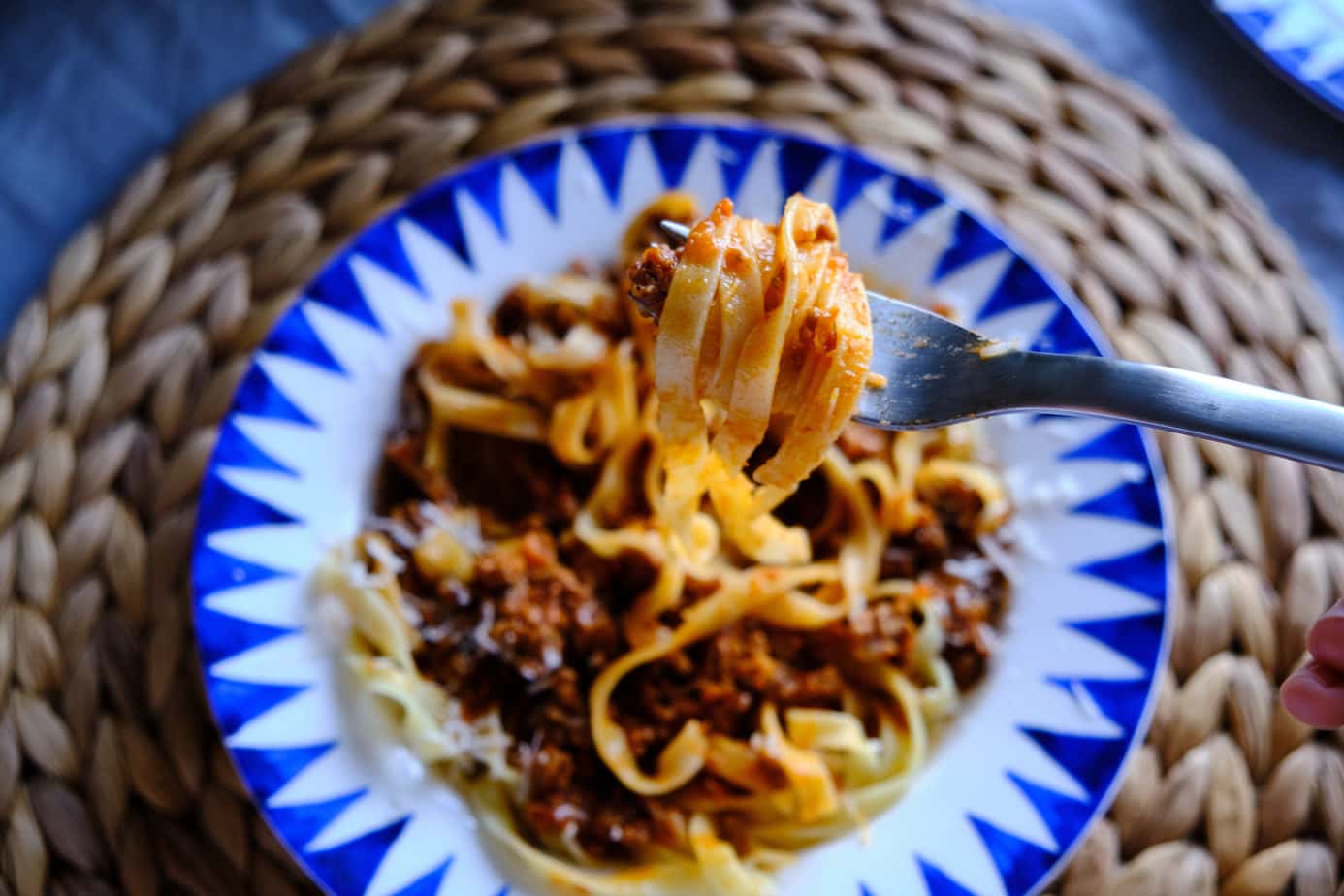 flat lay of a blue and white bowl on a circular seagrass placemat. The bowl is full of flat tagliatelle pasta and ground beef in a red ragu sauce. A fork is lifting some of the pasta up from the bowl towards the camera. Bologna Italy Food Guide.