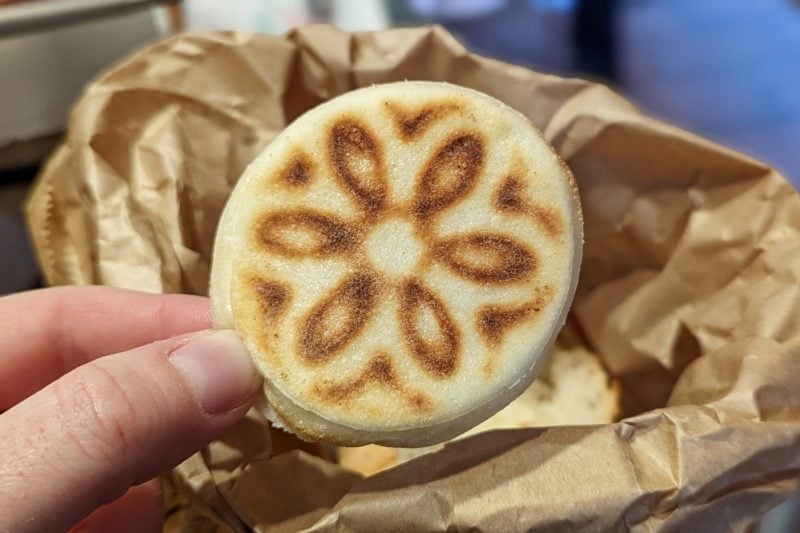 close up of a small round flat bread with a flower designed burned into it held by a thumb and forefinger in front of a brown paper bag