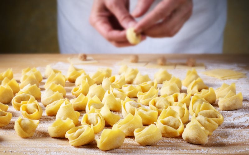 close up of a wooden board with lots of small filled pasta dumblings lightly dusted in flour. In the background a man's hands are shaping one of the pastas. 