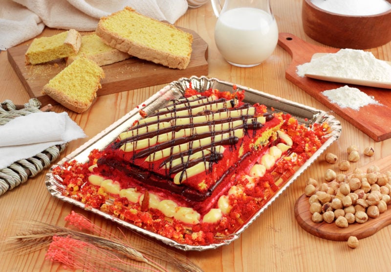 a large rectangular trifle with pink sponge and yellow custard lagers topped with a criss cross pattern of thick yellow custard and brown chcolate sauce stripes. the trifle is on a silver platter on a wooden table surrounded by various ingredients including nuts, sponge pieces, and a jug of milk. This is a traditional essert food in bologna italy. 