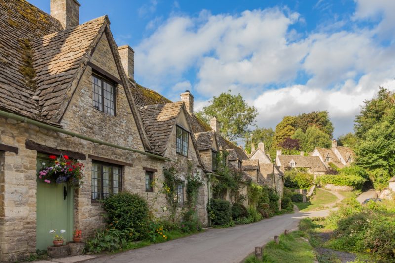 row of beige stone cottages in a line in front of a path next to grass and trees - cotswolds driving itinerary 