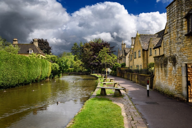 a wide river with green trees on either side and a path on the right next to some beige stone cottages