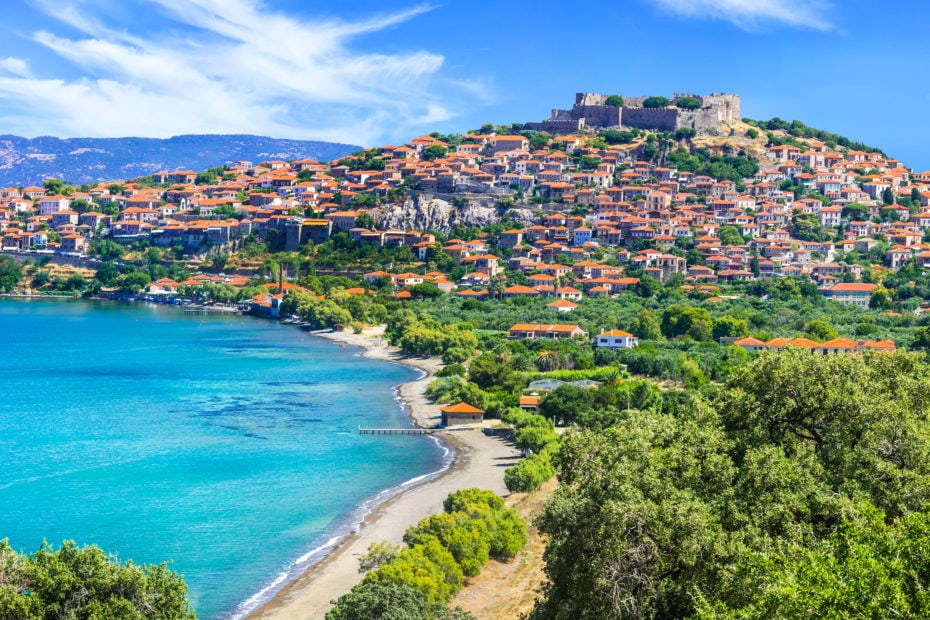 aerial view of Molyvos village in Lesvos with a curved bay and sandy beach next to turquoise water, the village is on the side of a hill beside the bay with a castle on top of the hill and red roofed buildings all around the side.
