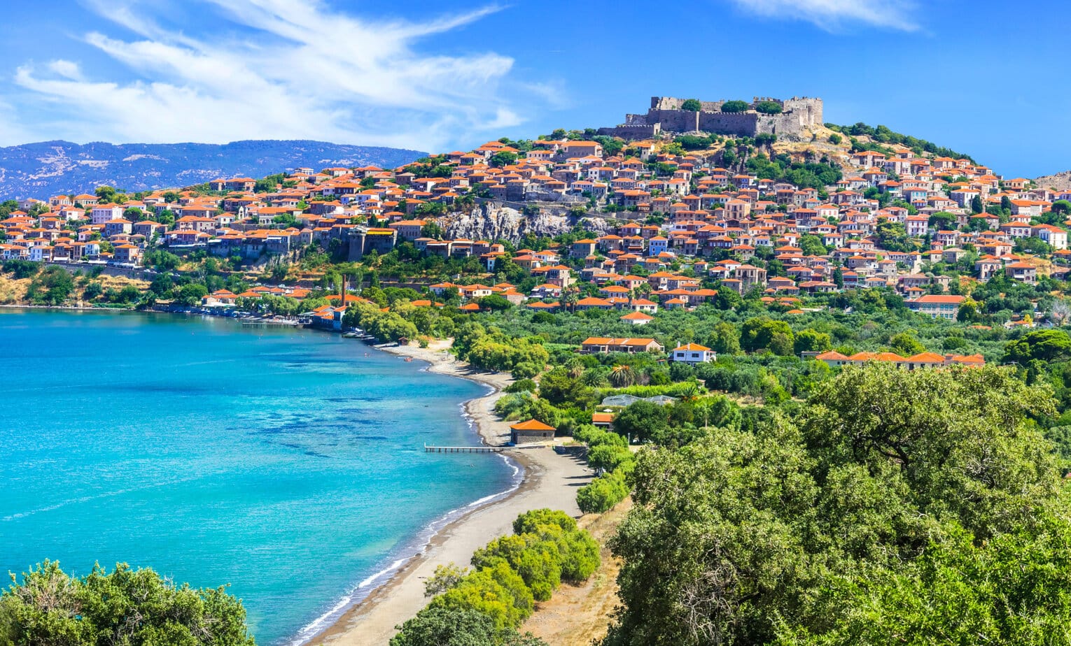 aerial view of Molyvos village in Lesvos with a curved bay and sandy beach next to turquoise water, the village is on the side of a hill beside the bay with a castle on top of the hill and red roofed buildings all around the side.