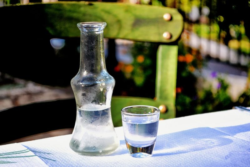 small glass bottle half full with clear spirit next to a small shot glass filled with the same spirit on an outdoor table with a white tablecloth with a green wooden chair out of focus behind