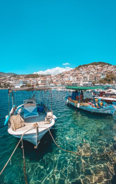 two small wooden fishing boats on turquoise water in Plomari harbour, the boat on the left is white with a grey awning over the deck and the boat on the right is blue and white with a green awning. there is a small town of whitewashed houses on the side of a hill in the background 