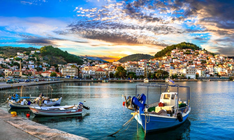 blue and white fishing boats in Plomari harbour on the island of Lesvos with three small hills and a town of whitewashed buildigns across the bay at sunset with a gold glow on the horizon above the hills. Things to do in Lesvos greece. 